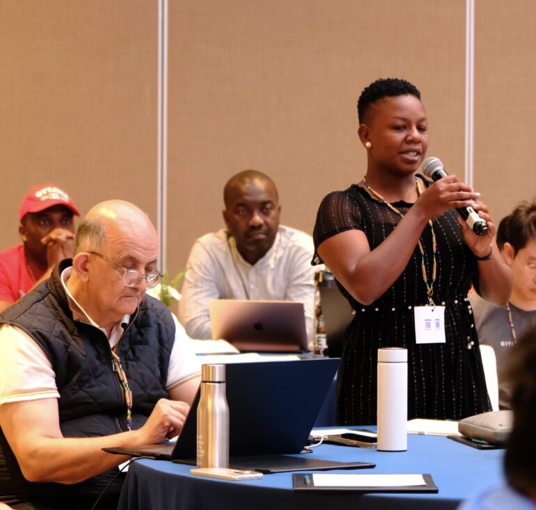 A woman stands and speaks into a microphone at a conference, while a man sits beside her typing on a laptop. Other attendees are seated in the background, listening and using laptops.
