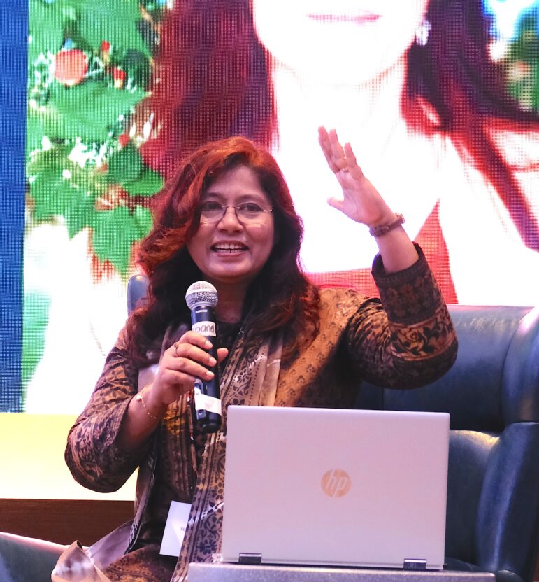 A woman with long dark hair and glasses speaks into a microphone and gestures with her hand while sitting in a chair behind an HP laptop. A colorful, blurred image is displayed in the background.