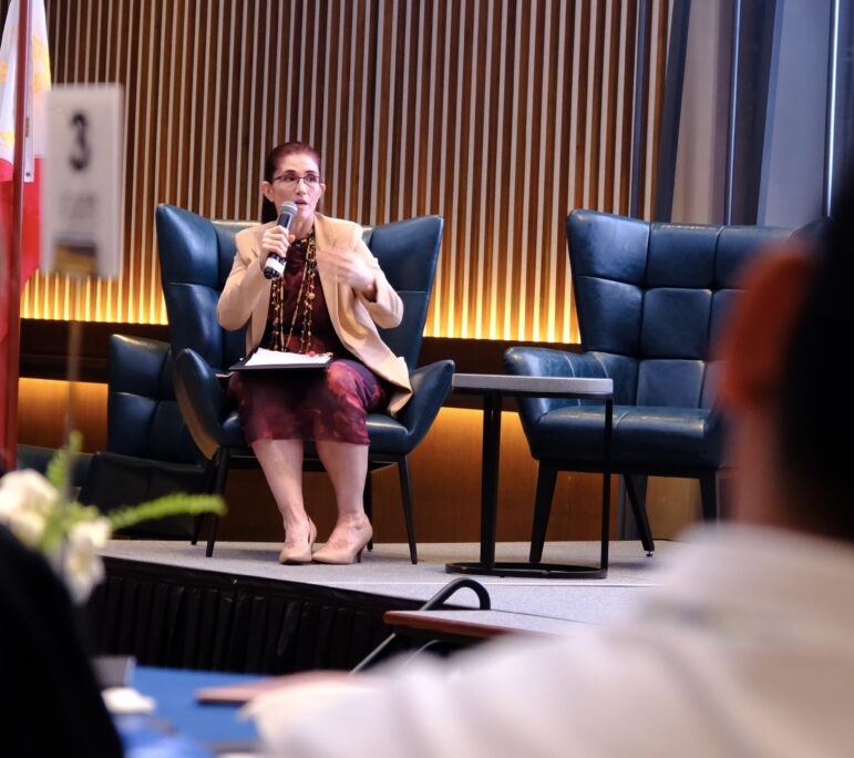 A person sits in a chair on stage, holding a microphone and speaking. They have short hair, glasses, and are wearing a tan blazer and patterned skirt. There is a table and an empty chair beside them.