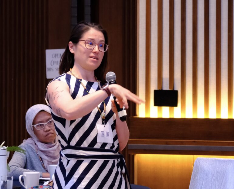 A woman wearing glasses and a black-and-white striped dress speaks into a microphone while gesturing with her hand. Another woman in a headscarf sits in the background, listening. The setting appears to be a conference or meeting.