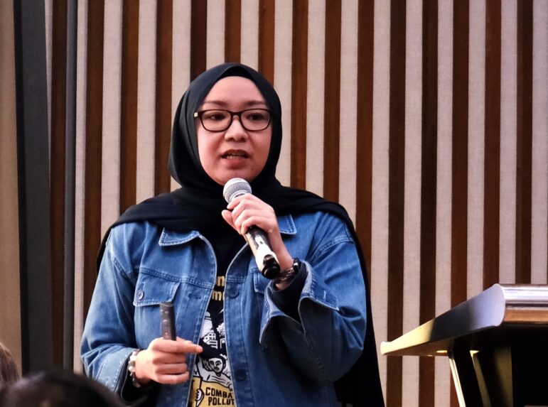 A person wearing glasses, a black hijab, and a denim jacket is speaking into a microphone while standing at a podium. Vertical wooden slats are visible in the background.