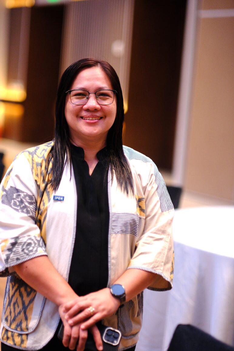 A woman with straight dark hair, glasses, and a patterned beige jacket stands indoors, smiling at the camera with her hands clasped. A round table and blurred background are visible.