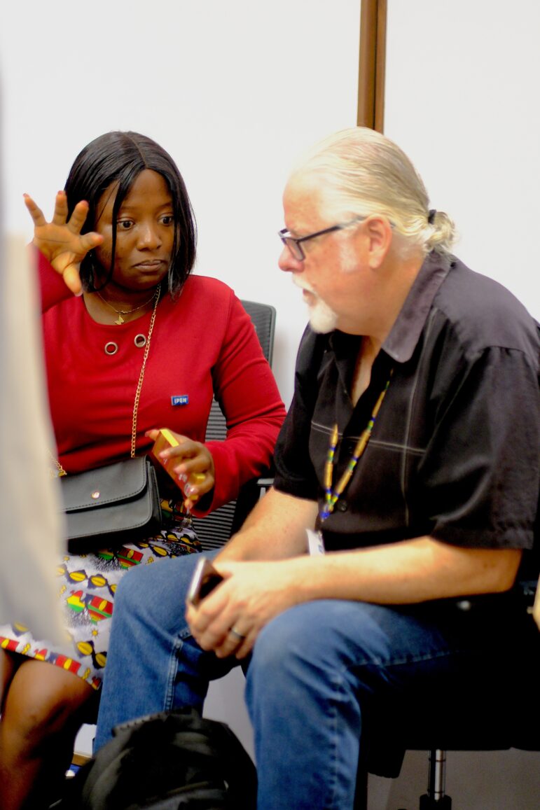 A woman in a red top gestures with her hand while speaking to a seated man with glasses and a white beard. Both appear focused on their conversation in an indoor setting.