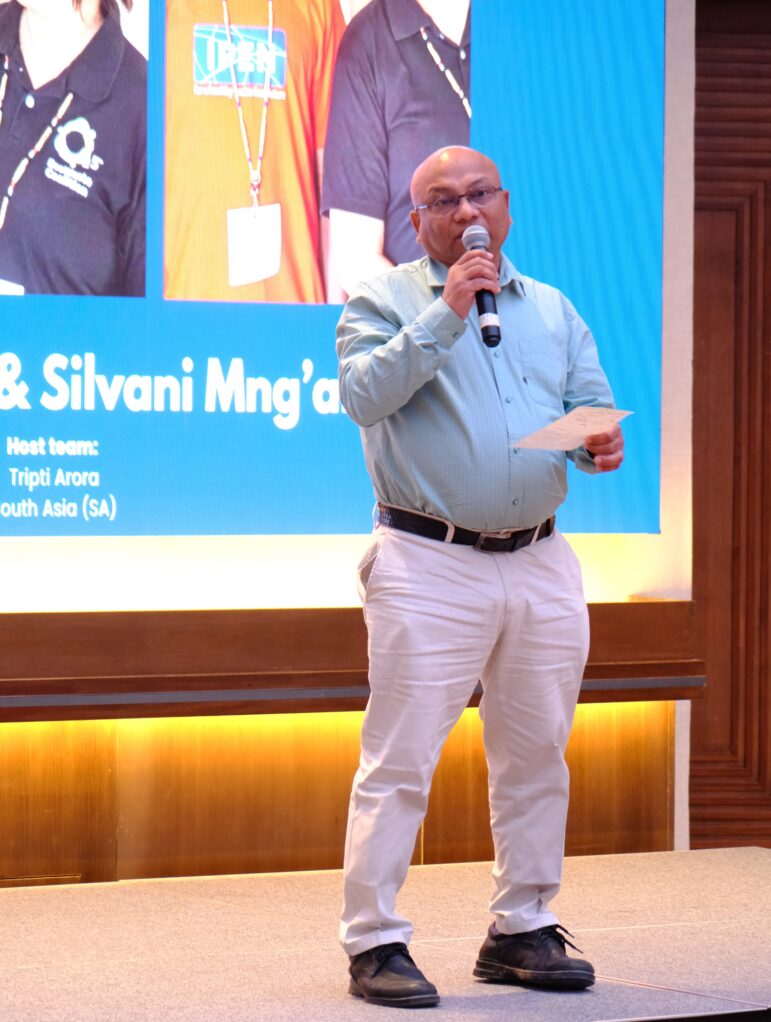 A bald man wearing glasses, a light blue shirt, white pants, and black shoes stands on stage holding a microphone and a piece of paper, speaking in front of a large screen displaying text and photos.