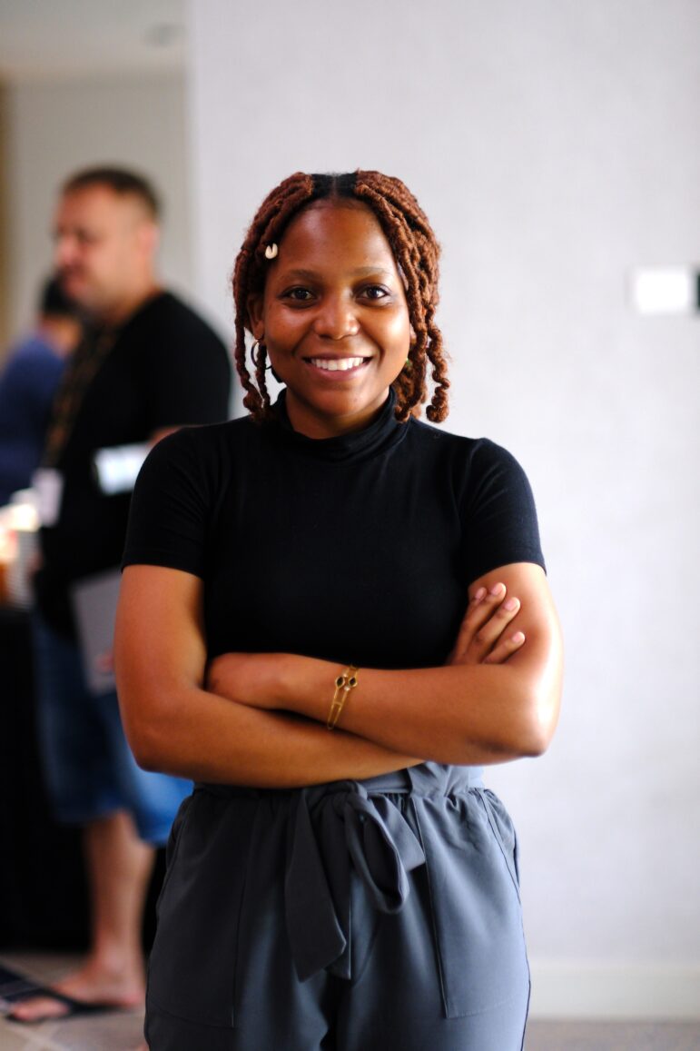 A woman with braided hair wearing a black short-sleeve shirt and gray pants stands indoors with her arms crossed, smiling at the camera. There are people and blurred background elements behind her.
