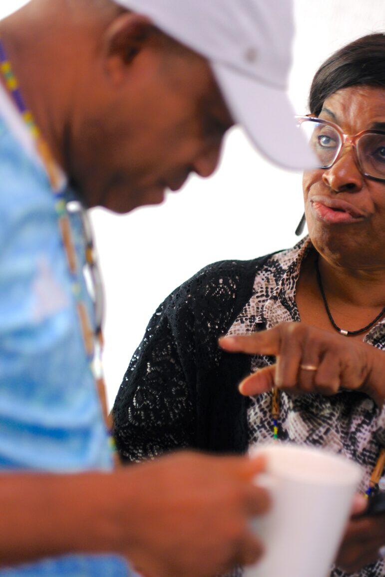A woman wearing glasses and a patterned top points and appears to speak to a man in a white hat and blue shirt, who holds a white cup. The background is brightly lit and out of focus.