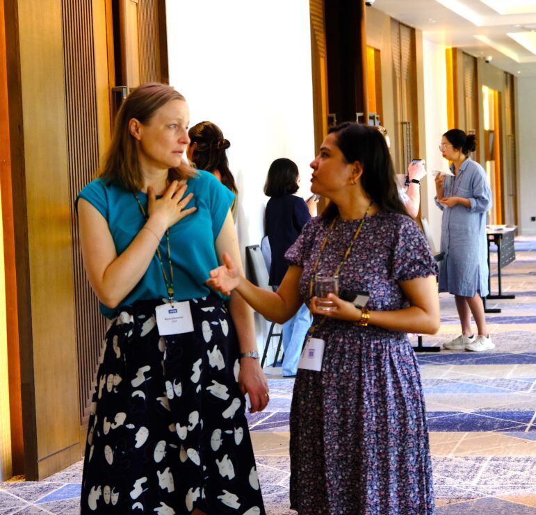Two women are standing in a hallway having a conversation, both wearing conference name badges. Other people are talking in the background. The setting appears to be a professional or networking event.