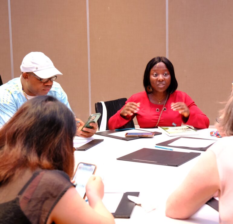 Four people sit around a white table engaged in discussion. One woman in red is speaking, while the others listen or look at their phones. Notebooks and phones are on the table, and the background is a plain beige wall.