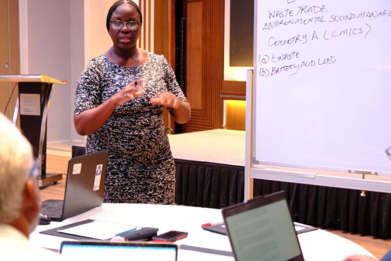 A woman stands near a whiteboard giving a presentation. The whiteboard has notes about waste trade and environmental concerns. Two laptops are open on the table in front of her, with people seated and listening.