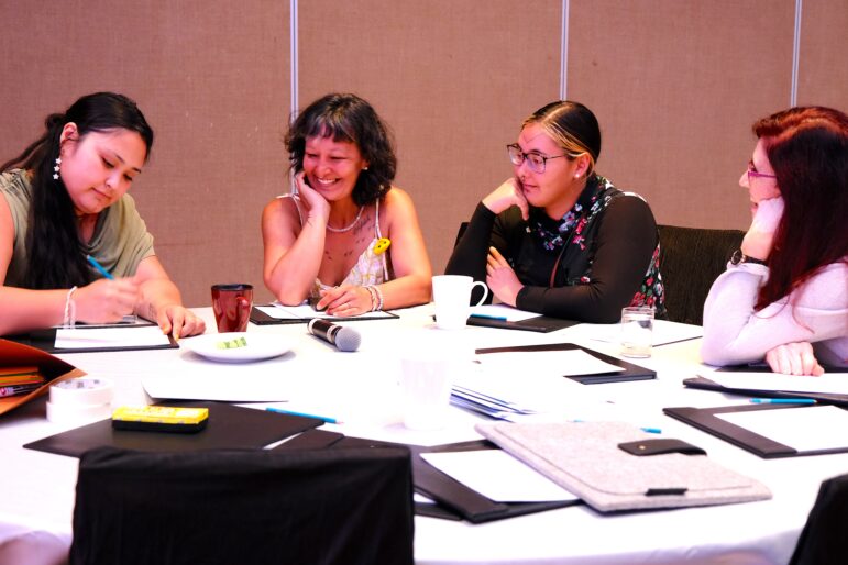 Four women sit around a round table with notebooks, drinks, and a microphone, engaged in discussion. One woman is writing while the others listen and watch her, appearing focused and attentive.