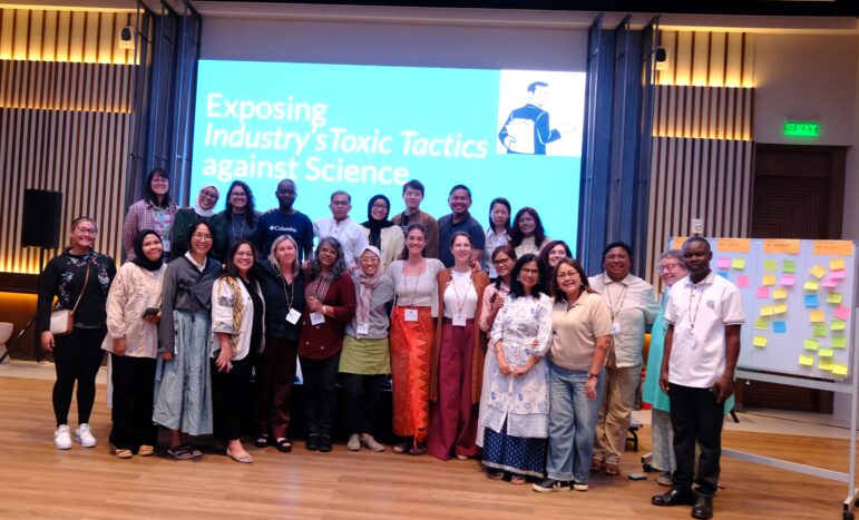 A group of around 25 people stand together and smile for a photo in a conference room. Behind them, a screen reads “Exposing Industrys Toxic Tactics against Science.” A board with sticky notes is visible on the right side.