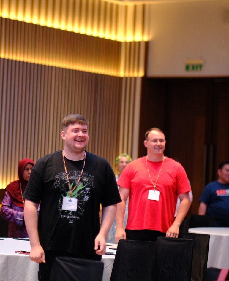 Several people stand and smile in a conference room. Two men in the front wear lanyards and name badges; one wears a black shirt, and the other a red shirt. Others and empty chairs are visible in the background.