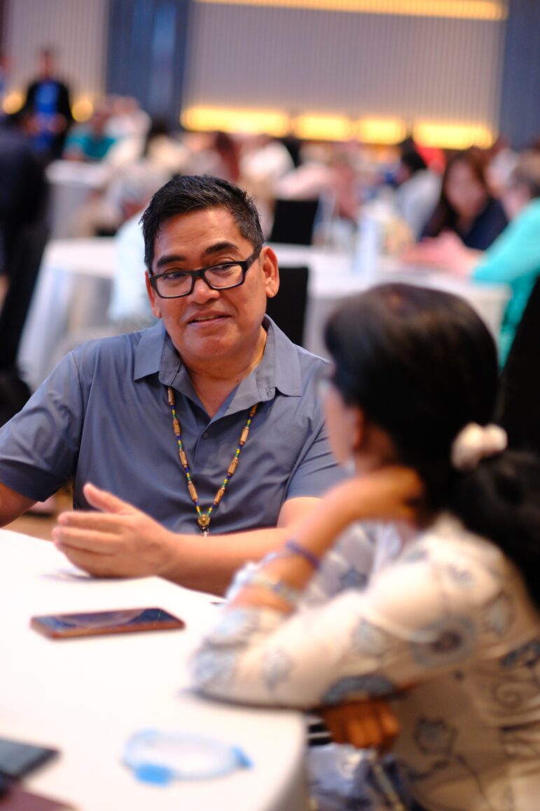 A man wearing glasses and a necklace is speaking to a woman across a table at a busy indoor event. Other people are visible in the background, seated at round tables.