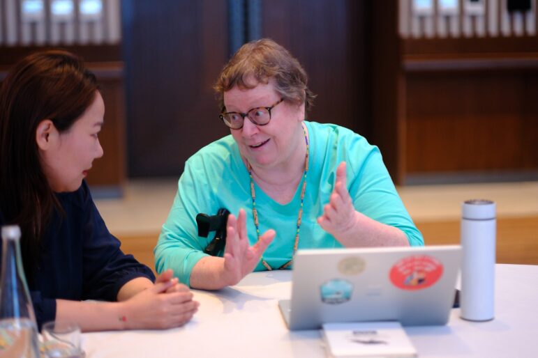 Two women sit at a table having a conversation. One woman gestures with her hands while speaking. A laptop with stickers, a notebook, and a bottle are on the table in front of them. The background is out of focus.