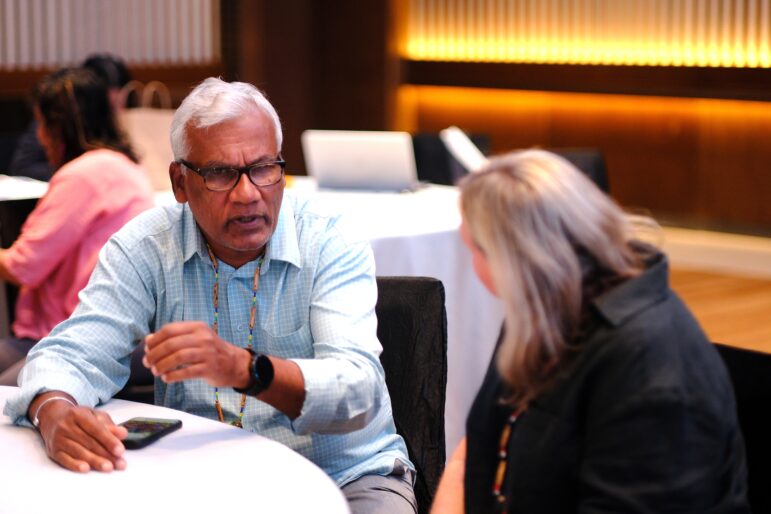 An older man wearing glasses and a blue shirt talks to a woman with blonde hair in a black jacket at a table in a conference room with blurred attendees in the background.