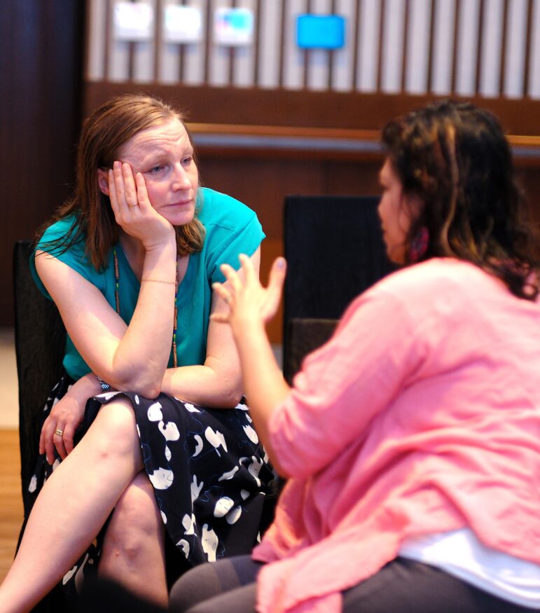 Two women are seated facing each other indoors; one woman in a green top listens with her head resting on her hand, while the other, in a pink top, gestures as she speaks.
