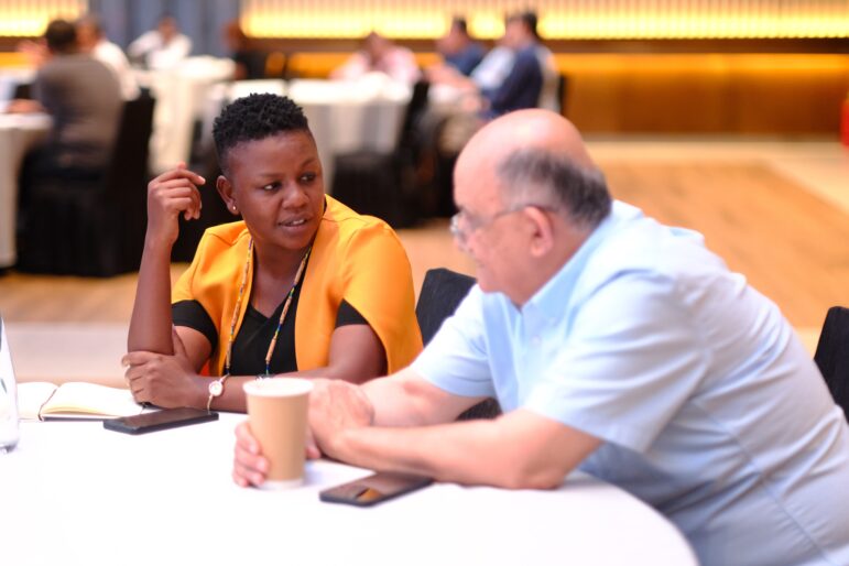 Two people sit at a round table having a conversation. One holds a paper cup, and the other has a notebook and pen. Both are dressed in business casual attire. Other people are visible in the blurred background.