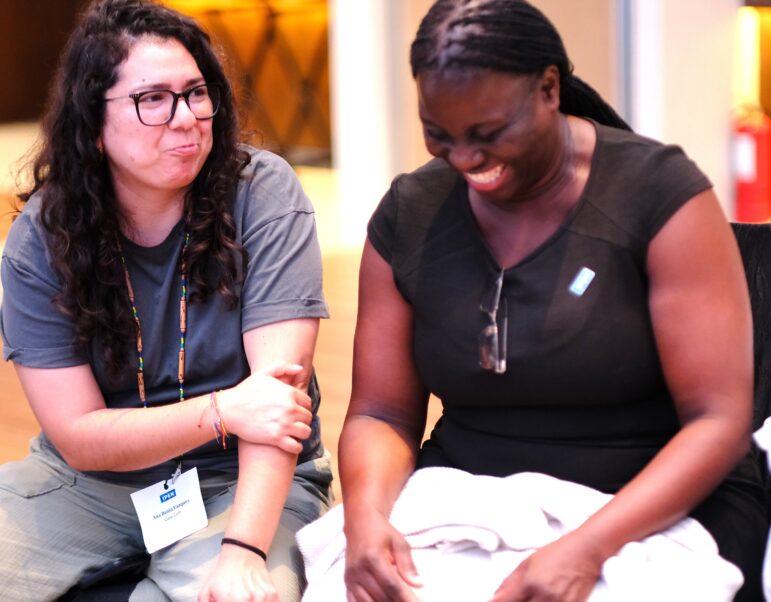 Two women sit side by side indoors, smiling and laughing together. One wears glasses and a name badge on a lanyard, while the other has glasses resting on her shirt and holds a light-colored cloth.