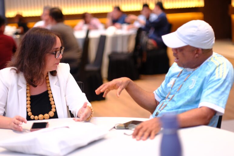A woman and a man sit at a round table engaged in conversation. The woman wears glasses, a white jacket, and a chunky necklace. The man wears a light blue jersey and a white cap. Other people are seated in the blurred background.