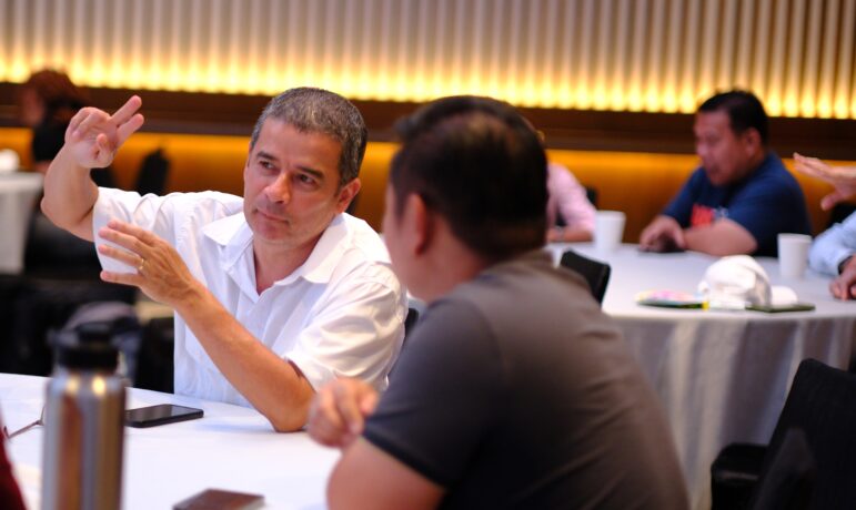 A man in a white shirt gestures while speaking to another man at a round table in a conference or meeting room, with several people in the background.