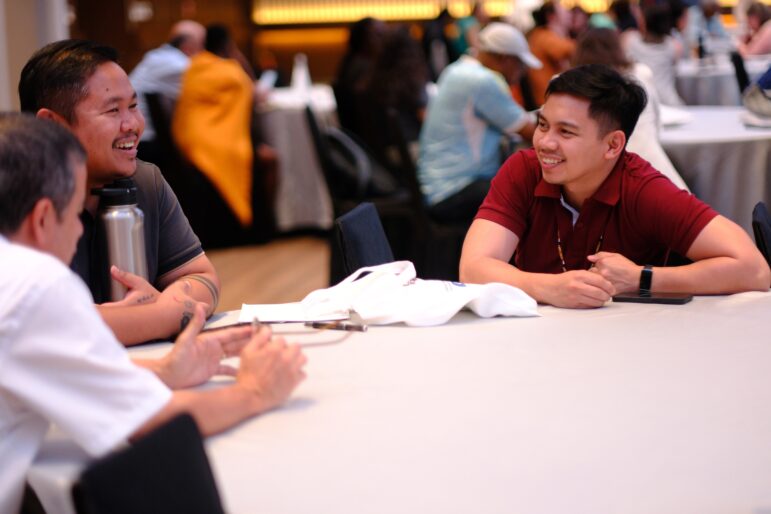 Three men sit at a round table engaged in conversation, smiling and appearing relaxed. Other groups of people are seated at tables in the background in a large indoor setting.