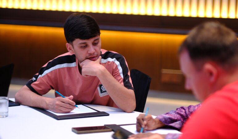 A man in a pink shirt sits at a table with a notebook and pen, appearing to think. Another person is writing beside him. There are a glass of water and a smartphone on the table. The background is softly lit.