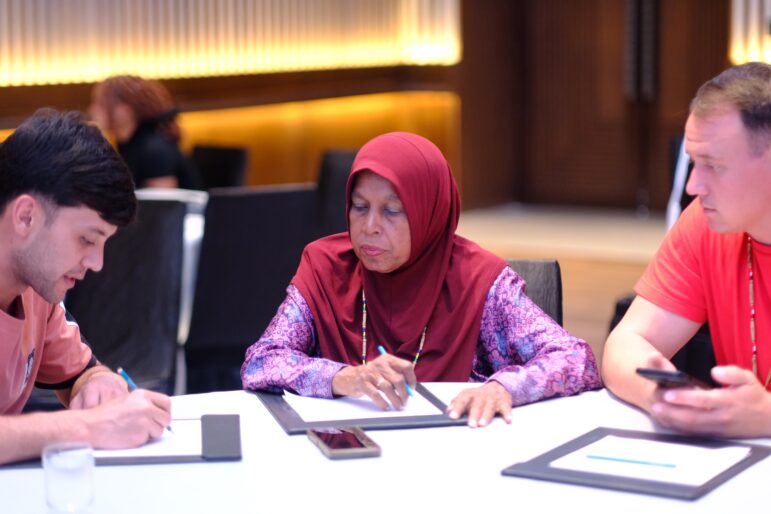 Three people sit around a table in a conference room, writing on notepads. A woman in a maroon hijab is in the center, with two men on either side. A phone and tablet are on the table.