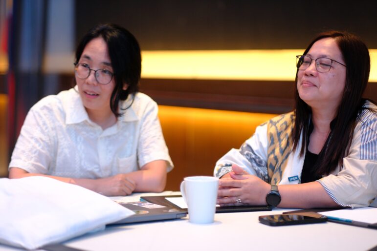 Two people with long hair and glasses sit at a table with papers, a folder, a smartphone, and a white mug in front of them. Both are looking slightly to the side, appearing attentive in a meeting or discussion.