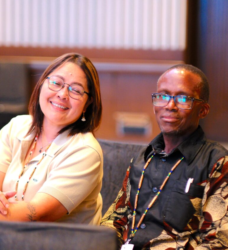Two people sit next to each other indoors, both wearing glasses and smiling at the camera. One person wears a light-colored shirt, and the other wears a patterned shirt. They both have conference lanyards around their necks.