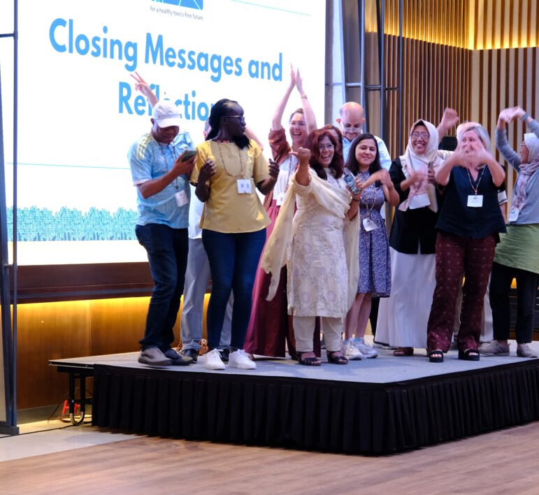 A group of people stand on a stage in front of a screen displaying the words Closing Messages and Reflection. They are smiling, waving, and posing together. Some people are wearing lanyards and name badges.