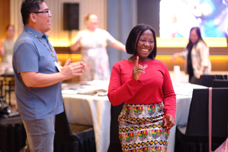 A woman in a red top and colorful skirt smiles and gestures with her finger while walking indoors, with people around her clapping and tables set up in the background.