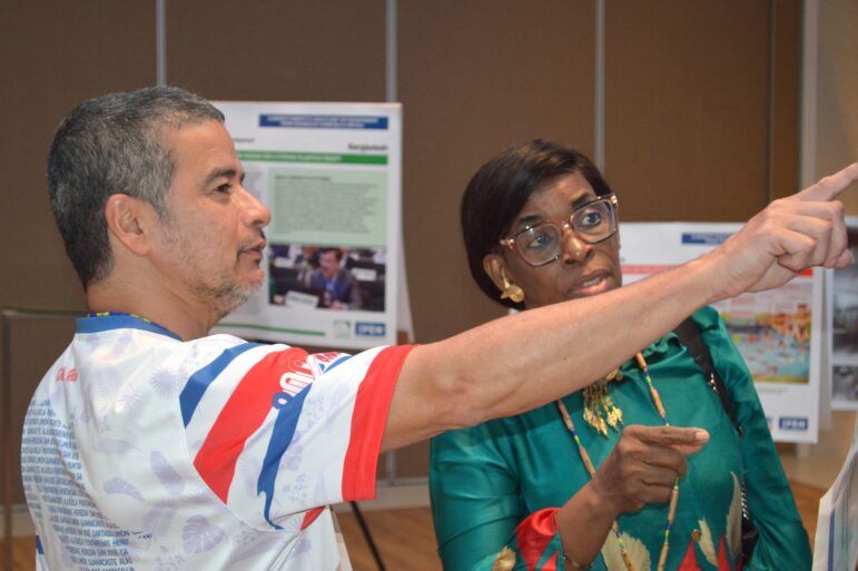 A man and a woman stand indoors near informational posters. The man is gesturing and pointing while speaking, and the woman, wearing glasses, looks in the direction he is pointing. Both appear engaged in conversation.