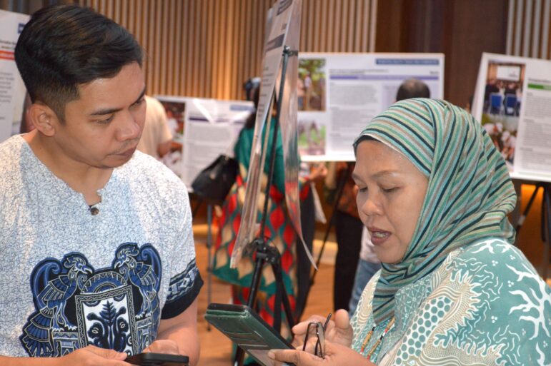 A man and a woman stand indoors, both holding mobile devices and having a conversation. Display boards with informational posters are visible in the background. The woman wears a striped headscarf.