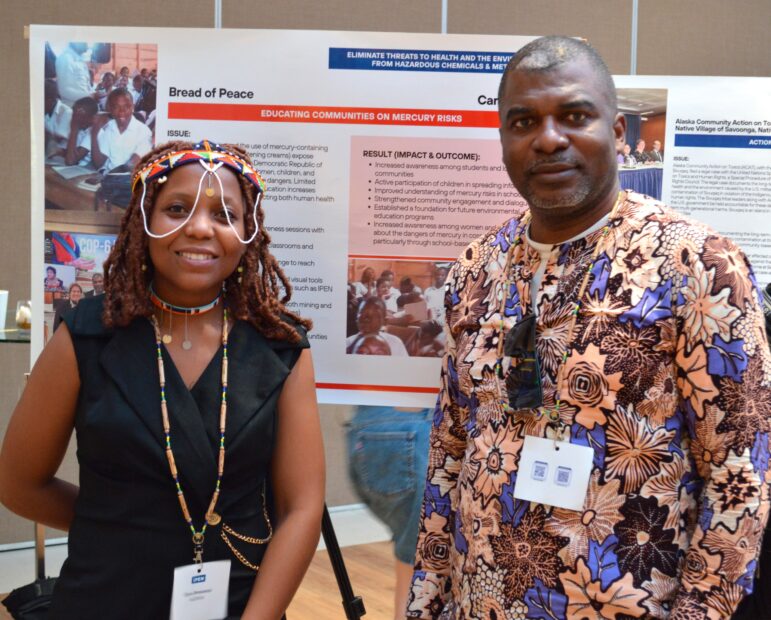 Two people stand indoors in front of a presentation poster about mercury risks and community health education. Both are wearing name badges; the woman is dressed in black with beaded jewelry, and the man wears a patterned shirt.