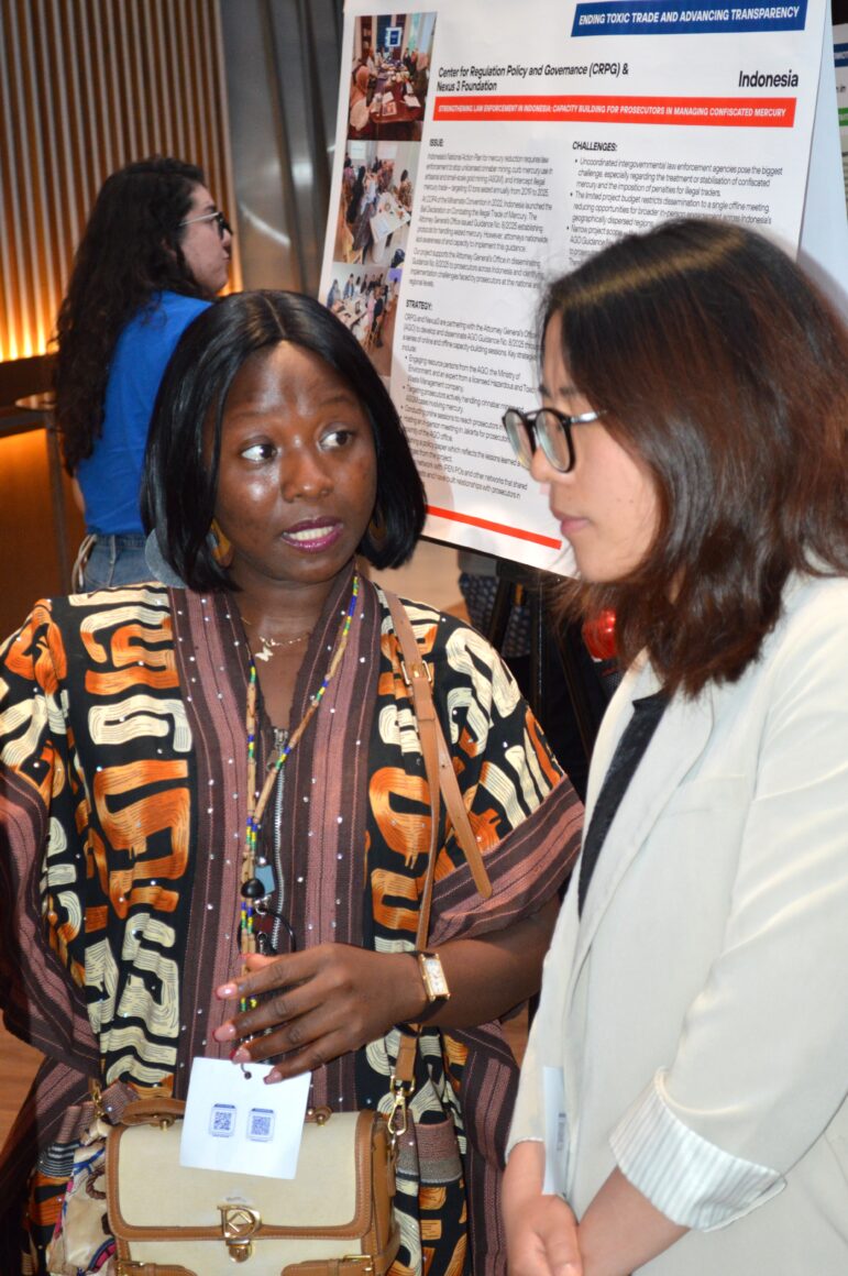 Two women stand and converse in front of a poster presentation about Indonesia’s trade and transparency policies at an indoor event. One woman wears a patterned outfit and the other a light-colored blazer.