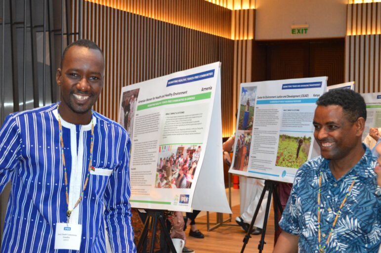 Two men stand and smile indoors in front of display boards with informational posters. The background shows vertical wooden wall panels and an exit sign above a door.