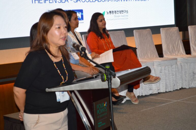 A woman speaks at a podium with a microphone while three other women sit on chairs in the background. A large screen displaying text is visible behind them.