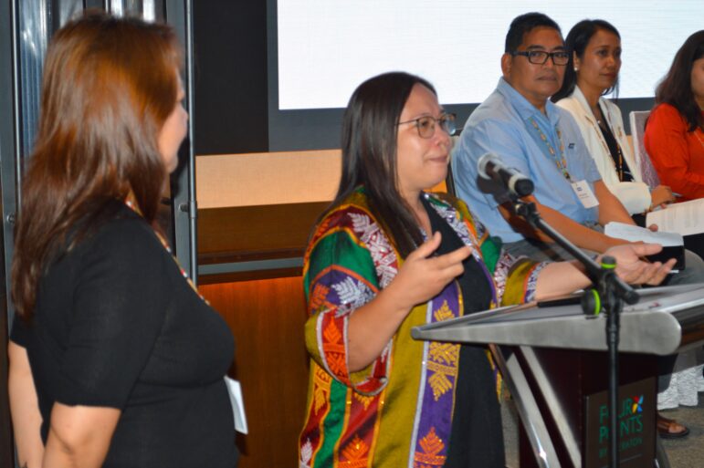 A woman wearing glasses and a colorful patterned jacket speaks at a podium with a microphone, while four other people stand nearby, three holding papers.