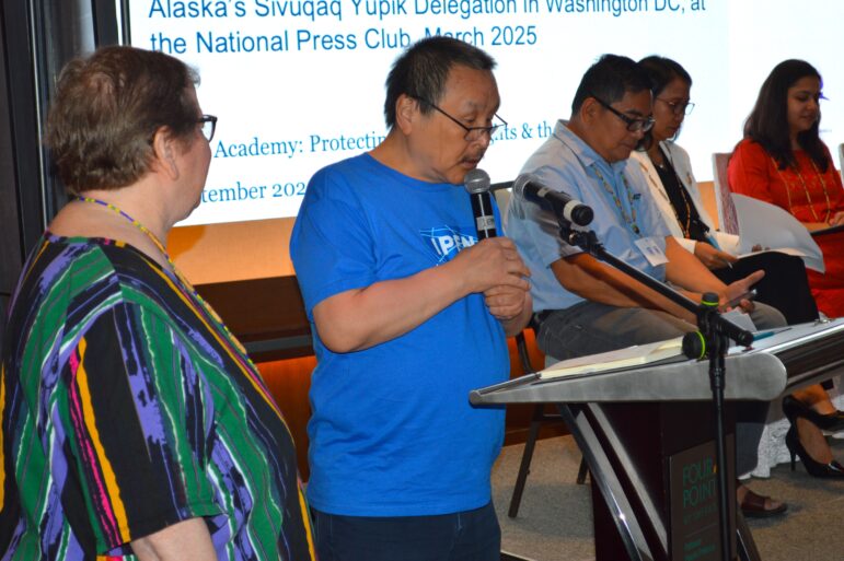 A man speaks into a microphone at a podium, while five people sit in a row behind him. A presentation slide with text is projected on a screen in the background.