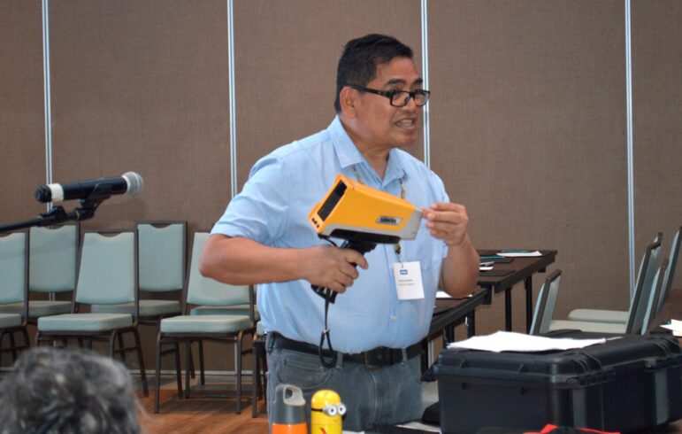 A man in glasses and a blue shirt holds a yellow handheld device while speaking in front of a group. A microphone, chairs, and a table with various items are visible in the background.