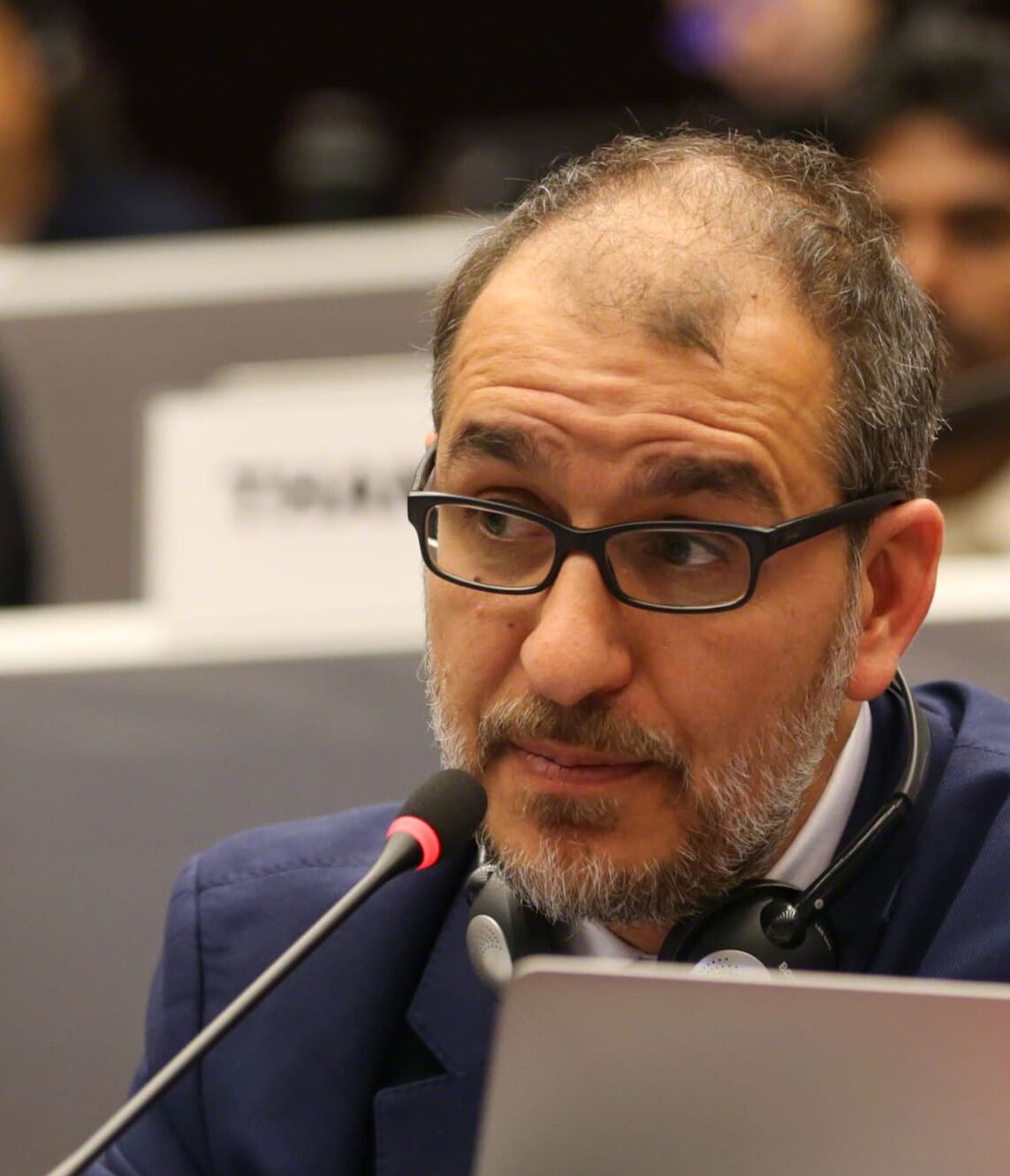 A man with glasses and a beard, wearing a dark suit and headphones around his neck, speaks into a microphone at a conference or meeting.