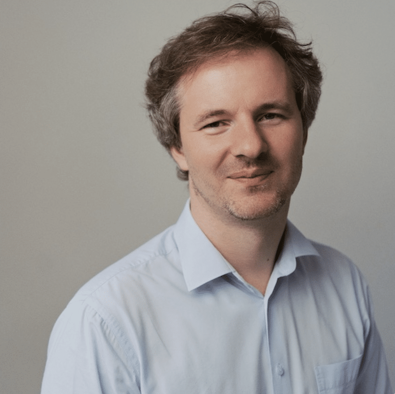 A man with light skin and short, slightly messy brown hair, wearing a light blue button-up shirt, stands in front of a plain light gray background and looks at the camera with a slight smile.