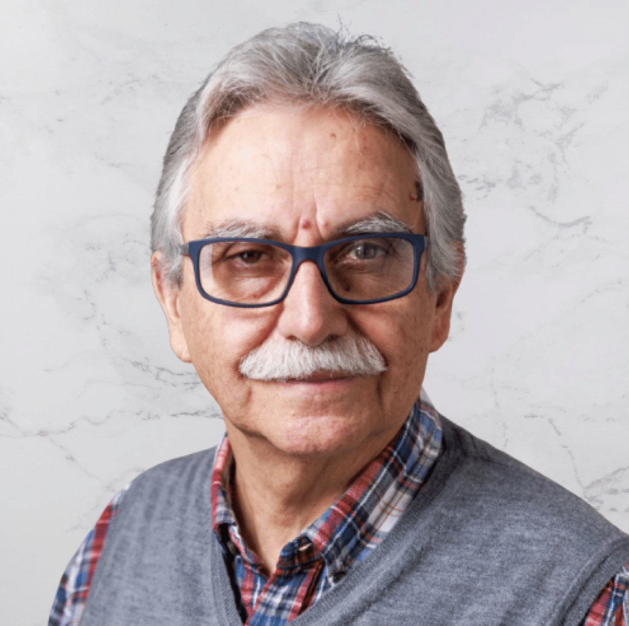 An older man with gray hair, a white mustache, and blue glasses, wearing a plaid shirt and a gray sweater vest, smiles slightly in front of a light-colored, marble-patterned background.