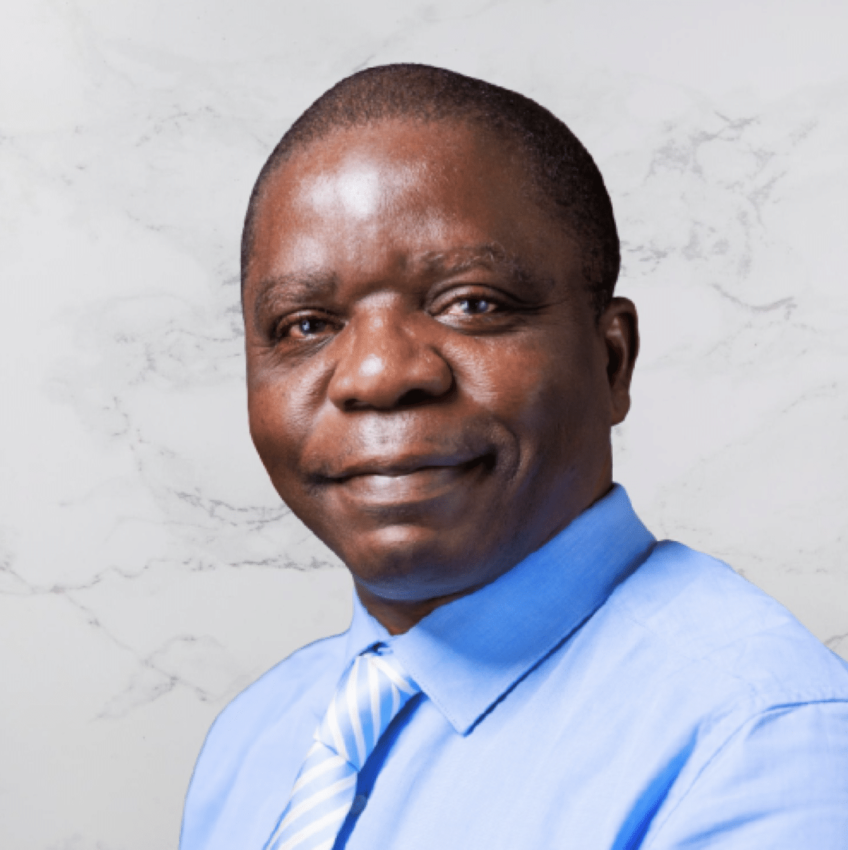 A man wearing a light blue dress shirt and a blue-and-white striped tie is smiling slightly while posing in front of a light marble background.