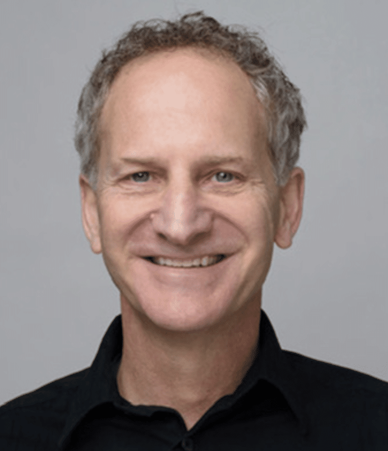 A middle-aged man with short, curly gray hair is smiling at the camera. He is wearing a black collared shirt and is posed in front of a plain light gray background.