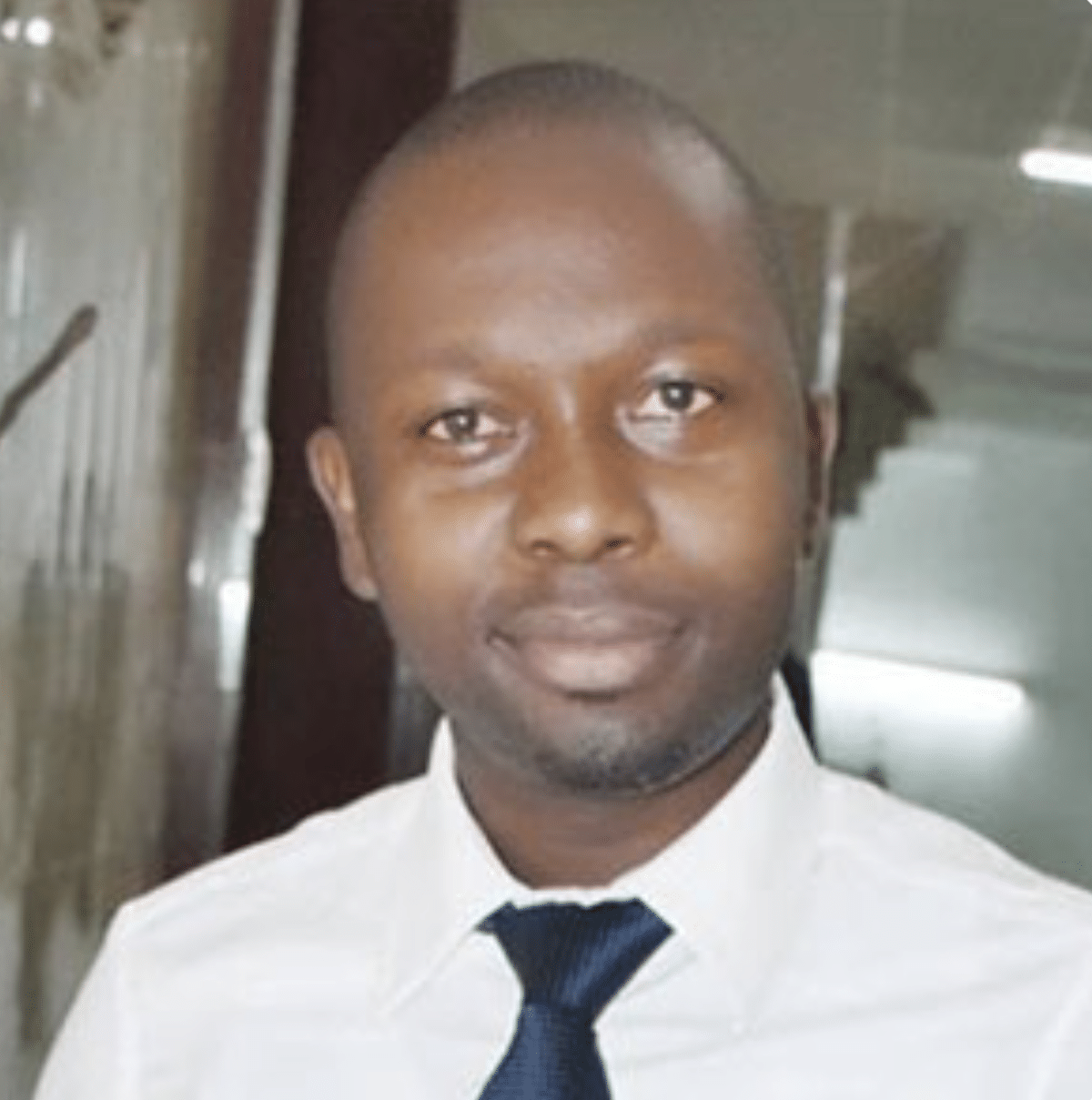 A man wearing a white dress shirt and a dark blue tie is standing indoors. He is facing the camera and has a neutral expression. There is a glass wall and some lights in the background.