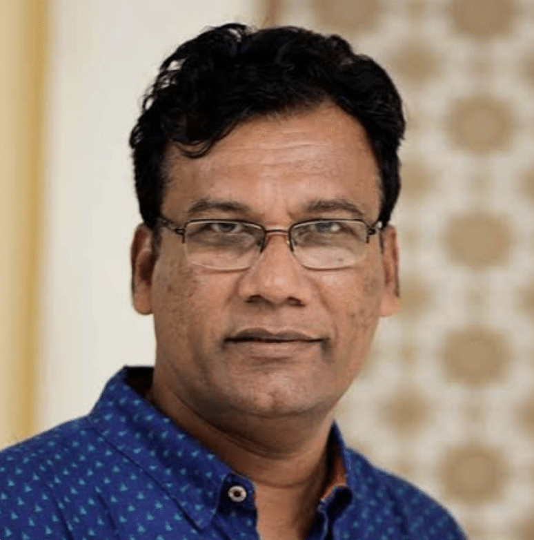 A man with short curly black hair, glasses, and a blue patterned shirt looks directly at the camera. The background is softly blurred with beige and white tones.