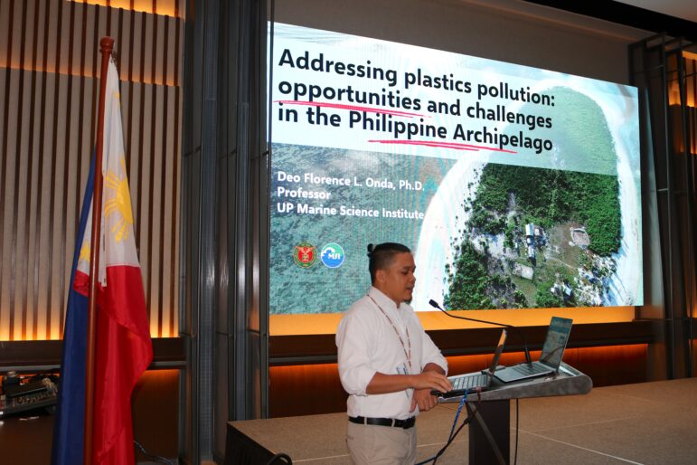 A man stands at a podium giving a presentation titled “Addressing plastics pollution: opportunities and challenges in the Philippine Archipelago.” A Philippine flag is next to him and a large screen displays his slides.