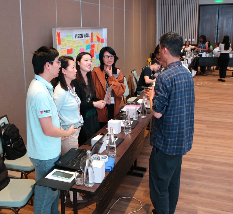 Five people stand by a table covered with laptops, glasses, and papers, having a discussion in a conference room. Colorful sticky notes are posted on the wall behind them. Other groups are visible in the background.