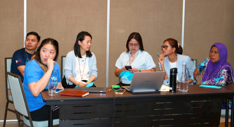 Six people sit at a table with laptops, notebooks, water bottles, and glasses. Some look at each other and talk, while others face forward. The background is a plain, brown wall.
